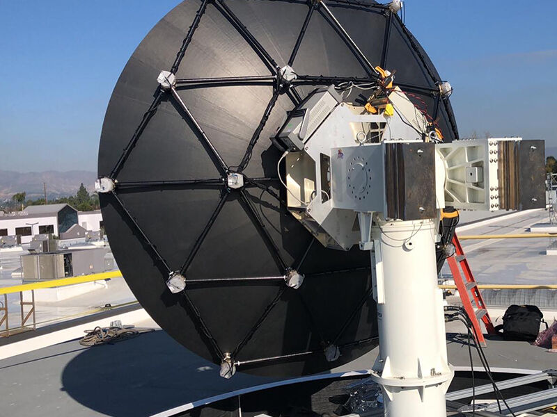 Megalodon dimensionally tunable reflector is deployed in the field. Hardware is on a rooftop, and there is a blue sky and mountains in the distance. The reflector is attached to a large column with electronics equipment and wiring visible. The image view is looking at the back of the reflector with the deformation grid.